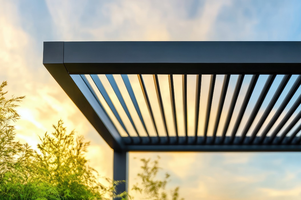 Eine moderne Pergola mit Lamellen wird vor einem lebhaften Sonnenuntergangshimmel mit umgebender Vegetation dargestellt. Harmonie zwischen Außenarchitektur und Natur